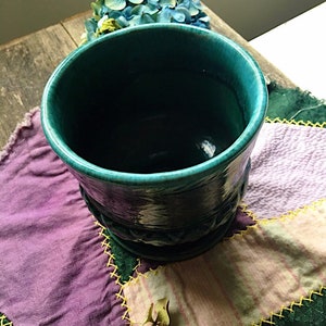 May include: A teal blue ceramic planter with a textured design. The planter is sitting on a patchwork quilt with purple, green, and pink fabric.