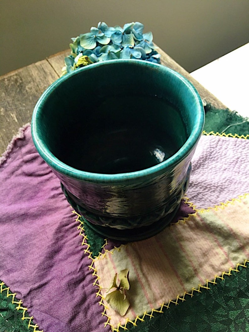 May include: A teal blue ceramic planter with a textured design. The planter is sitting on a patchwork quilt with purple, green, and pink fabric.