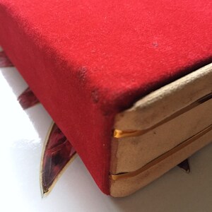 K&ouml;nnte beinhalten: Nahaufnahme einer roten Samtbox mit goldfarbenem Besatz. Die Box hat eine rechteckige Form und eine weiche, pl&uuml;schige Textur. Die Kanten sind mit goldfarbenem Metall verst&auml;rkt.