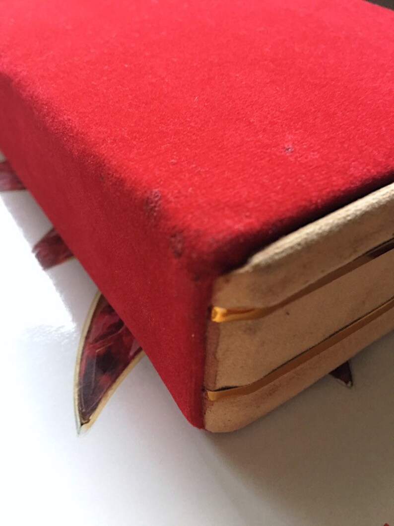 K&ouml;nnte beinhalten: Nahaufnahme einer roten Samtbox mit goldfarbenem Besatz. Die Box hat eine rechteckige Form und eine weiche, pl&uuml;schige Textur. Die Kanten sind mit goldfarbenem Metall verst&auml;rkt.