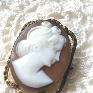 May include: An oval-shaped cameo brooch featuring a white carved profile of a person with curled hair set against a light brown background. The cameo is encased in a gold-toned metal frame with a textured, scalloped edge and a pin back.