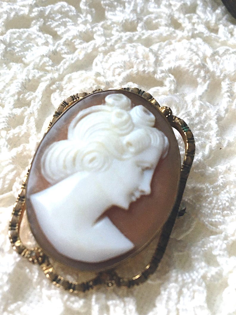 May include: An oval-shaped cameo brooch featuring a white carved profile of a person with curled hair set against a light brown background. The cameo is encased in a gold-toned metal frame with a textured, scalloped edge and a pin back.