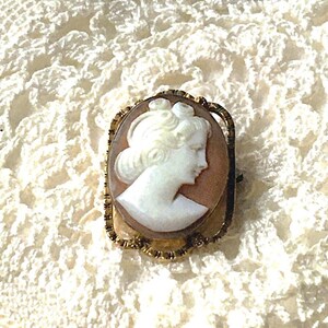 May include: An oval-shaped brooch featuring a white carved cameo profile of a person set against a light brown background. The cameo is encased in a textured gold-tone metal frame with a scalloped edge and a pin mechanism on the back.