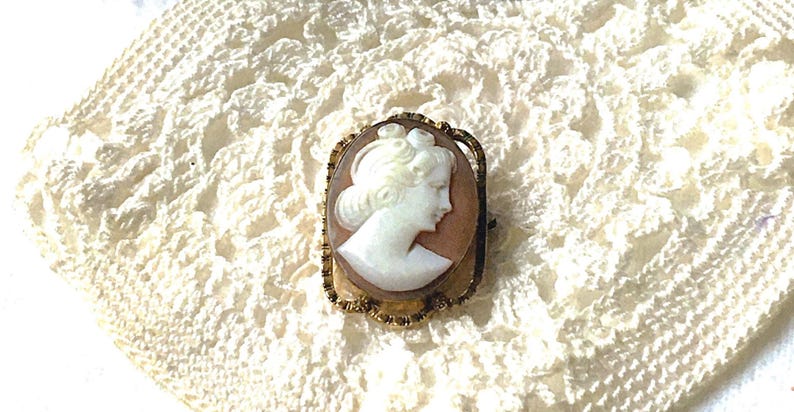 May include: An oval-shaped brooch featuring a white carved cameo profile of a person set against a light brown background. The cameo is encased in a textured gold-tone metal frame with a scalloped edge and a pin mechanism on the back.