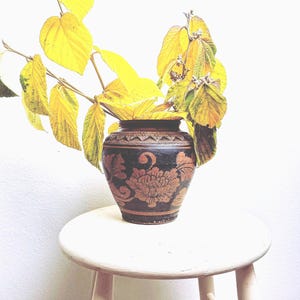 May include: A black and brown decorative vase with a floral design, filled with yellow leaves. The vase sits on a small, round, light-colored wooden stool. The leaves have a vibrant yellow hue, contrasting with the dark vase.