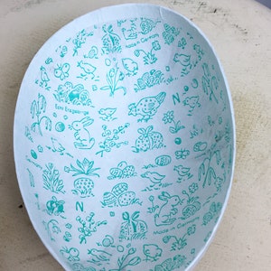 May include: A white paper Easter egg with a blue and green pattern of bunnies, chicks, and eggs. The text "Echt Erzgebirge" and "Made in Germany" are printed on the egg.