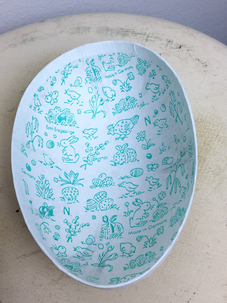 May include: A white paper Easter egg with a blue and green pattern of bunnies, chicks, and eggs. The text "Echt Erzgebirge" and "Made in Germany" are printed on the egg.