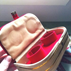 K&ouml;nnte beinhalten: Ge&ouml;ffnete Schmuckschatulle mit rotem Samtinnenraum und zwei Ringhaltern. Die Au&szlig;enseite ist beige mit goldenen Zierleisten und einem roten Verschluss. Das Innenfutter ist hellbeige.