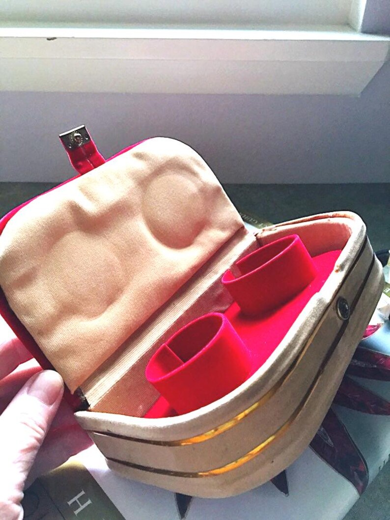 K&ouml;nnte beinhalten: Ge&ouml;ffnete Schmuckschatulle mit rotem Samtinnenraum und zwei Ringhaltern. Die Au&szlig;enseite ist beige mit goldenen Zierleisten und einem roten Verschluss. Das Innenfutter ist hellbeige.