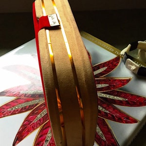 K&ouml;nnte beinhalten: Eine rechteckige Schmuckschatulle in Beige mit goldenen Streifen und rotem Samtinnenraum. Ein kleines goldenes Etikett ist an der Schatulle befestigt. Die Schatulle liegt auf einer dekorativen Oberfl&auml;che mit einem rot-wei&szlig;en Blumendesign.