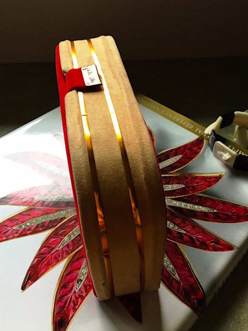 K&ouml;nnte beinhalten: Eine rechteckige Schmuckschatulle in Beige mit goldenen Streifen und rotem Samtinnenraum. Ein kleines goldenes Etikett ist an der Schatulle befestigt. Die Schatulle liegt auf einer dekorativen Oberfl&auml;che mit einem rot-wei&szlig;en Blumendesign.
