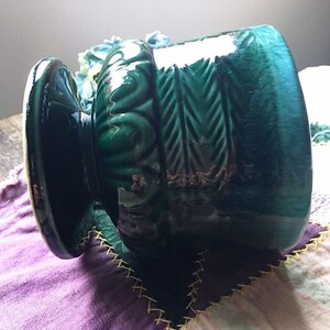 May include: A green ceramic planter with a textured design and a wide base. The planter has a glossy finish and is decorated with a repeating pattern of leaves and arrows.