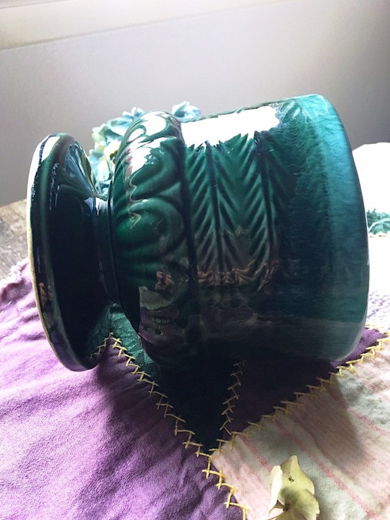 May include: A green ceramic planter with a textured design and a wide base. The planter has a glossy finish and is decorated with a repeating pattern of leaves and arrows.