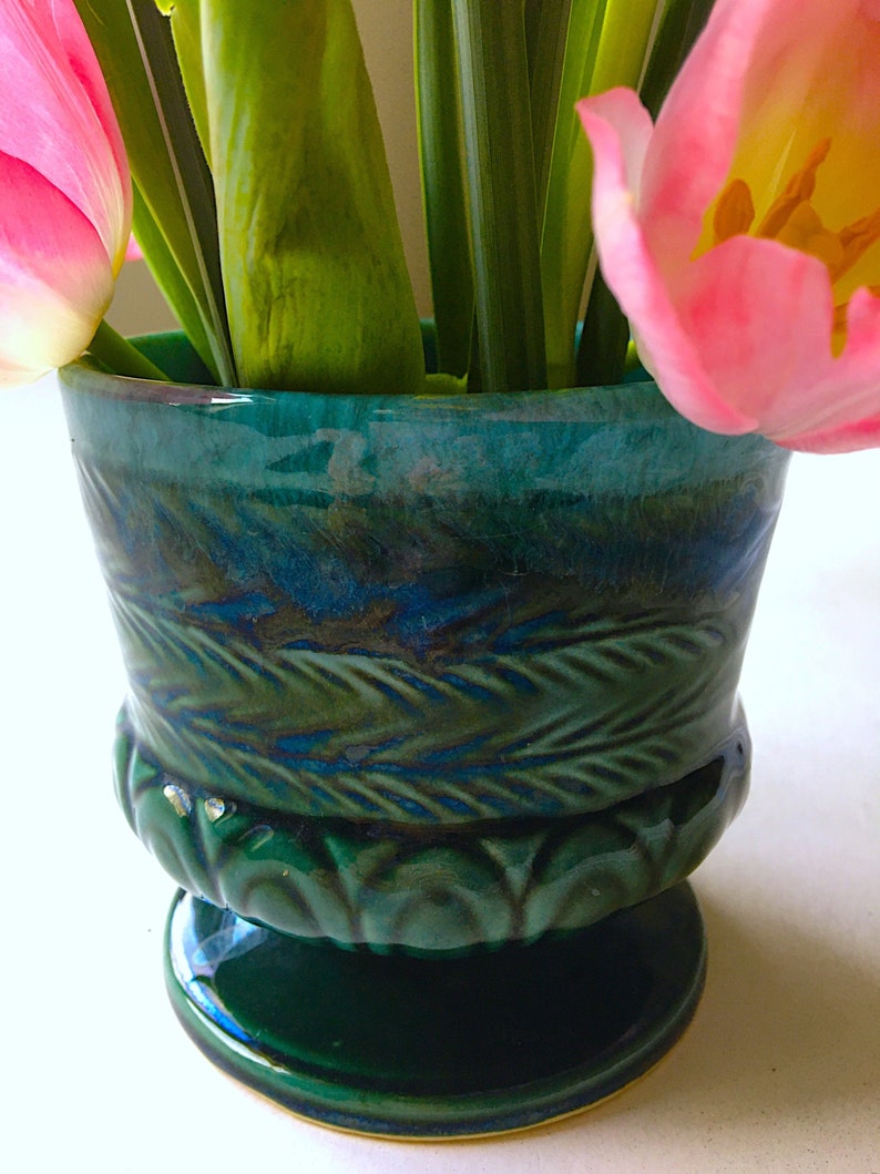 May include: A green ceramic vase with a textured design and a wide base. The vase is filled with pink tulips and green leaves.