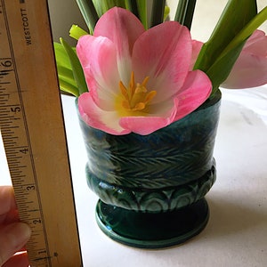 May include: A pink and white tulip with yellow center in a green ceramic vase with a leafy design. The vase has a scalloped base.