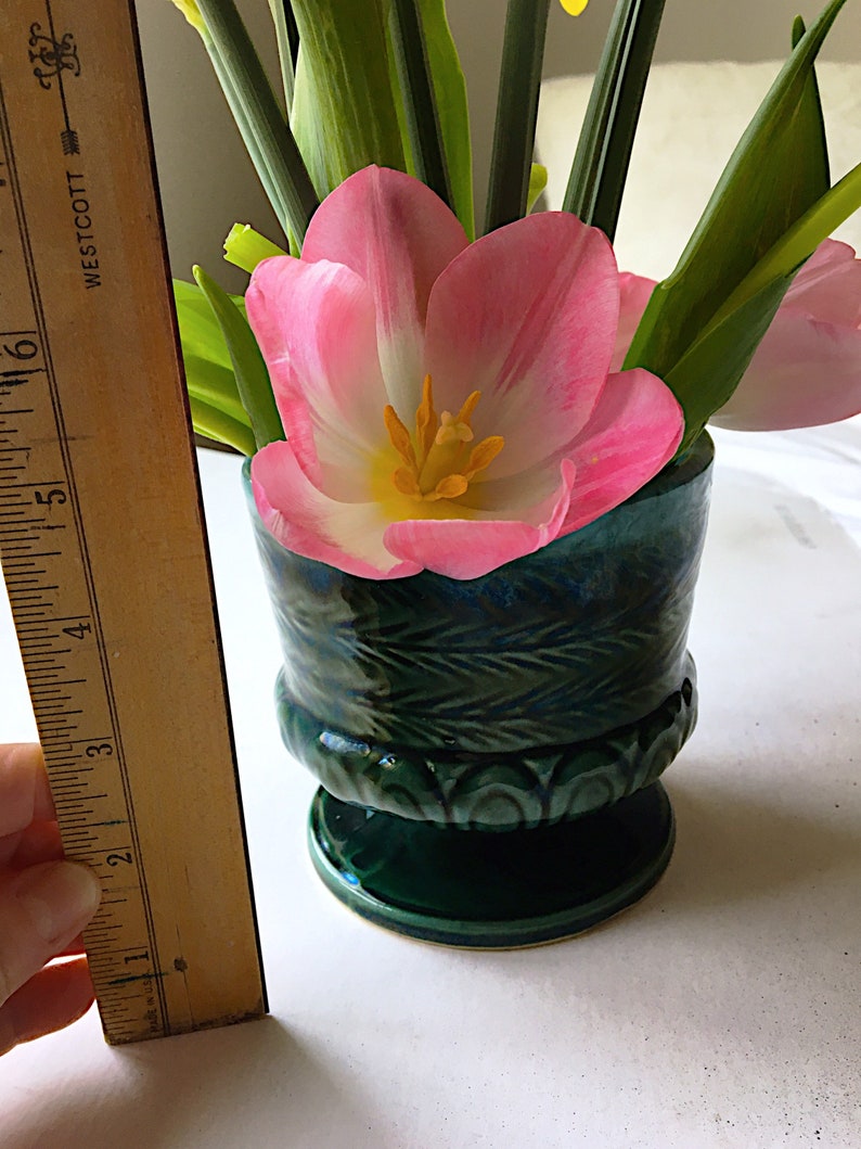 May include: A pink and white tulip with yellow center in a green ceramic vase with a leafy design. The vase has a scalloped base.