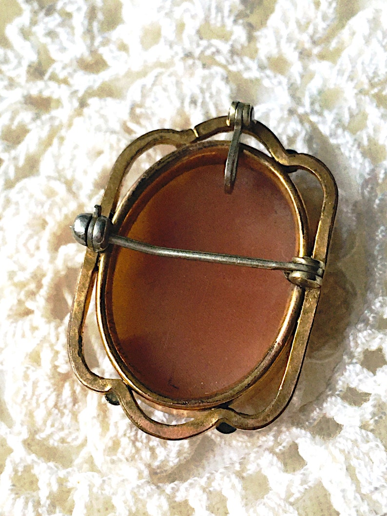 May include: An oval-shaped brooch featuring a smooth, light brown center set within a gold-toned metal frame. The back includes a horizontal pin mechanism with a safety clasp and a small vertical loop at the top for potential pendant use.