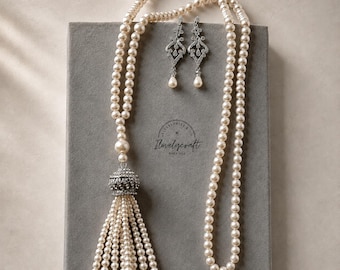 Peut inclure: Un long collier composé de perles couleur crème et d'un pompon surmonté d'une pièce argentée incrustée de cristaux transparents, accompagné de boucles d'oreilles assorties. L'ensemble est présenté sur une surface grise avec le texte Ilovelycraft.