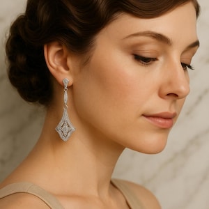 May include: A silver-toned drop earring featuring a geometric diamond-shaped pendant with intricate filigree metalwork and small embedded clear stones. The earring hangs from a small triangular top piece attached to a thin vertical metal bar.