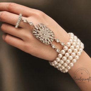 May include: A slave bracelet featuring a five-strand white pearl bracelet connected by two crystal-encrusted chains to a central oval medallion with a sunburst design, which attaches to a matching crystal-encrusted ring on the middle finger.