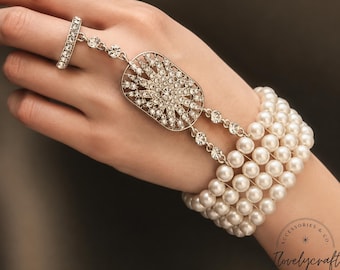 Op de afbeelding: Een slavenarmband met een vijfstrengs witte parelarmband verbonden door twee met kristallen ingelegde kettingen aan een centraal ovaal medaillon met een zonnestraalontwerp, dat vastzit aan een bijpassende met kristallen ingelegde ring.