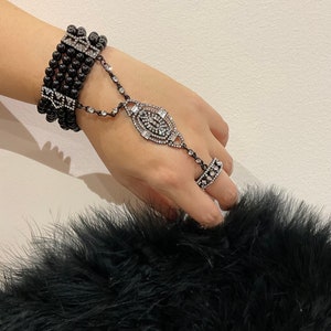 Puede incluir: Un brazalete negro de cuentas con un adorno de diamantes de imitaci&oacute;n y un anillo a juego. El brazalete est&aacute; conectado al anillo por una cadena.