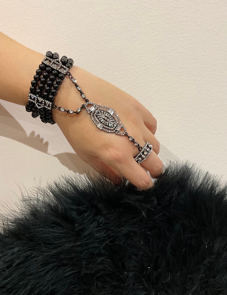 Puede incluir: Un brazalete negro de cuentas con un adorno de diamantes de imitaci&oacute;n y un anillo a juego. El brazalete est&aacute; conectado al anillo por una cadena.