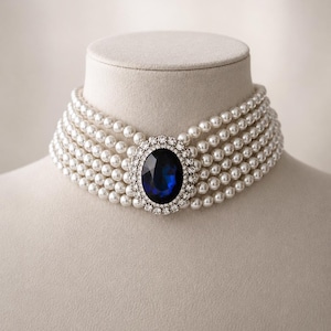 Peut inclure: Un collier ras-du-cou multi-rangs composé de cinq rangées de perles blanches. Le centre du collier est orné d'une grande pierre précieuse ovale bleu profond, entourée d'une bordure de petits cristaux clairs et étincelants.
