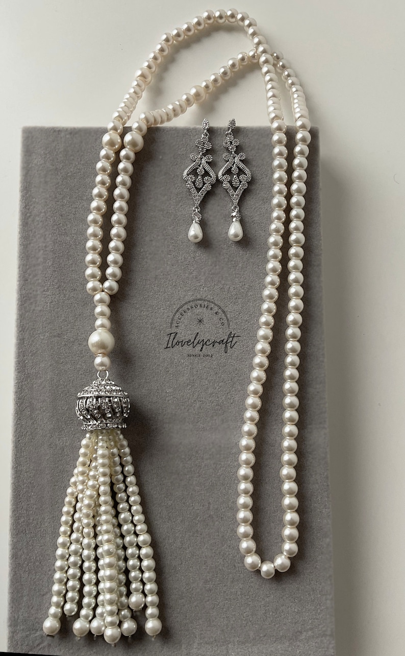 May include: A white pearl necklace with a crystal tassel pendant and matching crystal earrings. The necklace is on a gray background.
