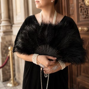 May include: A black feather fan with a dark, intricate handle held in front of a black velvet garment. The ensemble is accessorized with multiple strands of white pearl necklaces and matching multi-strand pearl bracelets featuring ornate silver clasps.