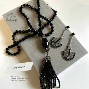 Puede incluir: Un collar negro con borla con un colgante de &oacute;nix negro y pendientes art d&eacute;co a juego. El collar est&aacute; hecho de cuentas negras y los pendientes son de plata con detalles negros.