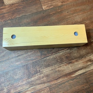 Puede incluir: Una caja de madera clara con dos agujeros en la parte superior. La caja est&aacute; sobre un suelo de madera.
