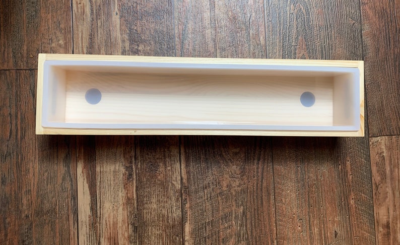 Puede incluir: Un molde de jab&oacute;n de madera con un inserto de silicona blanco. El molde tiene dos agujeros redondos en el inserto de silicona.