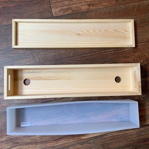 Puede incluir: Tres cajas de madera, una con dos agujeros, y un molde de silicona transparente. Las cajas son de madera clara y tienen forma rectangular. El molde es flexible y tiene una forma ligeramente curvada.