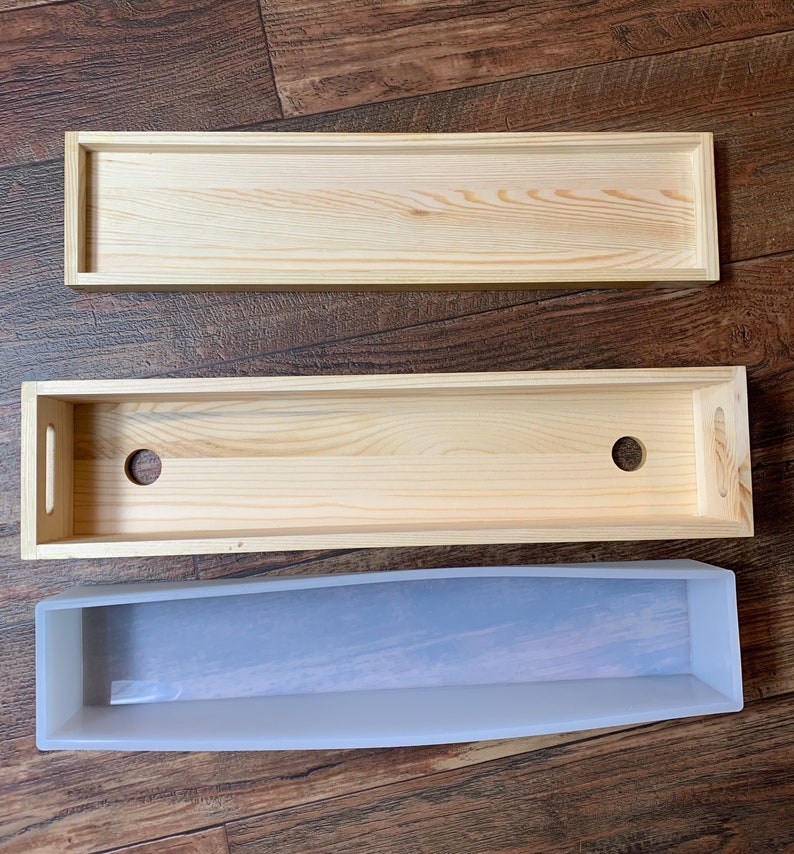 Puede incluir: Tres cajas de madera, una con dos agujeros, y un molde de silicona transparente. Las cajas son de madera clara y tienen forma rectangular. El molde es flexible y tiene una forma ligeramente curvada.