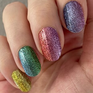May include: Close-up of a hand with four fingernails painted with different glittery nail polishes. The colours are yellow, green, pink, and purple.