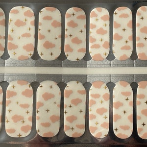May include: Nail art strips with a repeating pattern of pale pink clouds and gold stars on a white background. The strips are arranged in rows, ready for application. A whimsical design for a manicure.