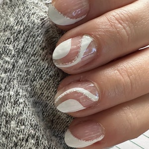 May include: Close-up of a hand with manicured nails. The nails feature a clear base with white and silver designs. Some nails have a white curved design with a silver glitter accent, while others have a white tip with a silver glitter line.