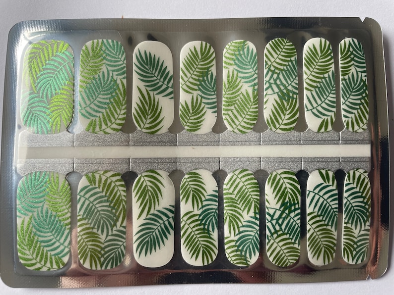 Puede incluir: Tiras de arte de u&ntilde;as con un dise&ntilde;o de hojas tropicales. Las tiras de u&ntilde;as presentan un patr&oacute;n repetido de hojas de palma verdes sobre un fondo blanco. Las tiras de u&ntilde;as est&aacute;n en un paquete plateado.