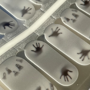May include: A sheet of nail polish wraps with a Halloween theme. The wraps feature black silhouettes of ghosts and hands on a white background.