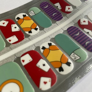Pode incluir: Uma folha de películas para unhas com tema de Alice no País das Maravilhas. As películas incluem imagens de cartas de baralho, um relógio de bolso e um padrão listrado.