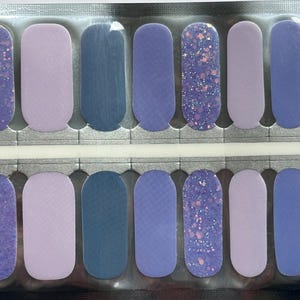 May include: A sheet of nail polish strips in various shades of purple and blue. The strips include solid colors, textured designs, and glitter accents. The nail art strips are arranged in a grid pattern, ready for application.