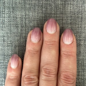 Puede incluir: Primer plano de una mano con cinco uñas pintadas con un efecto degradado. Las uñas hacen la transición de una base blanca a un tono púrpura claro. Las uñas, de forma ovalada, parecen bien cuidadas.