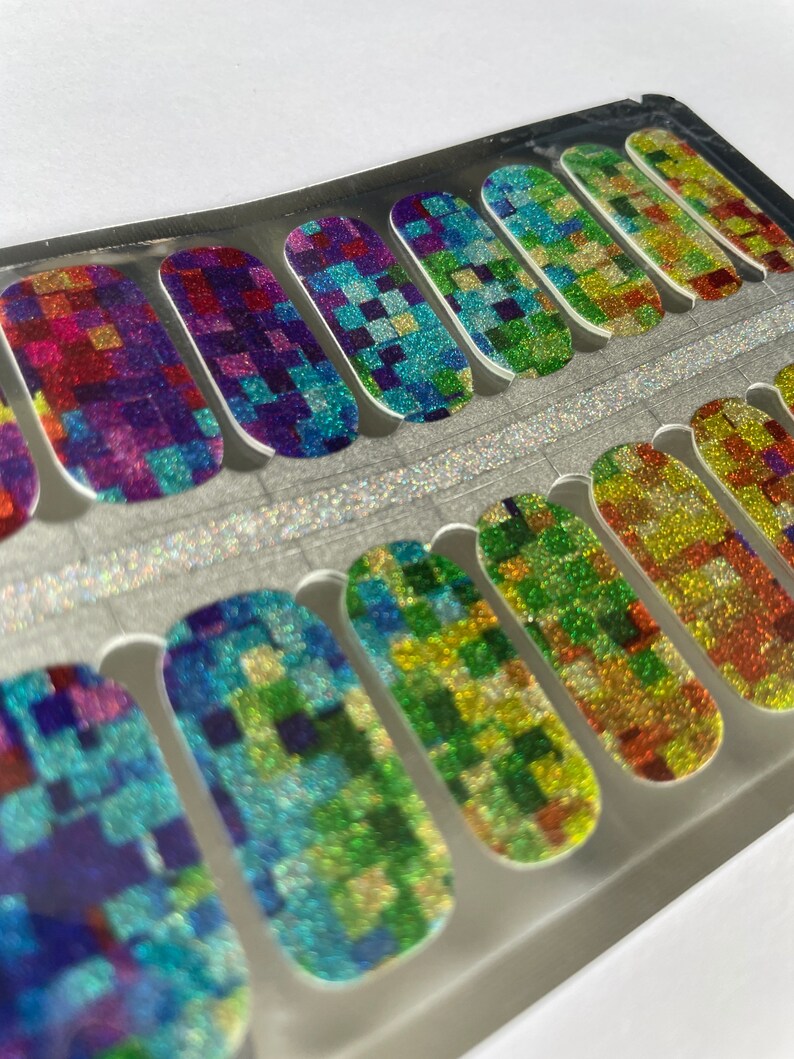 May include: A set of nail polish strips with a colorful, pixelated design. The strips feature a glittery finish and a variety of colors, including red, purple, blue, green, and yellow. The nail art is arranged in a clear plastic package.