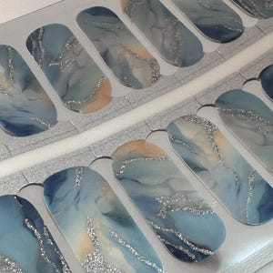 Puede incluir: Tiras de arte de uñas con un diseño veteado en tonos azules, grises y melocotón, acentuadas con purpurina plateada. Las tiras están dispuestas en filas sobre un respaldo transparente, mostrando una estética moderna y elegante. Estas tiras ofrecen una manicura de calidad de salón en casa.