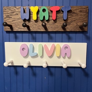 May include: Two wooden coat racks with hooks, one stained brown and one white. The top rack has the name "WYATT" spelled out in colorful letters. The bottom rack has the name "OLIVIA" spelled out in pastel colors.