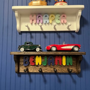 May include: Two wooden shelves with decorative items and names. The top shelf is white with the name "HARPER" in pastel letters. The bottom shelf is brown with the name "BENJAMIN" in colorful letters. Toy cars are on the bottom shelf.
