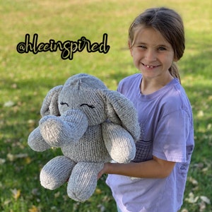 May include: A young person holding a gray knitted elephant plush toy. The toy has a sweet expression with closed eyes and a small smile. The plush toy is soft and cuddly, perfect for a child's bedroom.