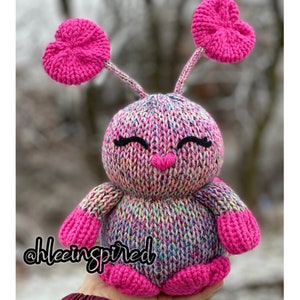 May include: A pink knitted toy with heart-shaped ears and a colorful, speckled body. The toy has a smiling face with black eyelashes and a pink nose. The toy is held in a hand with the text "@hleeinspired" visible in the bottom left corner.