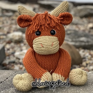 May include: A crocheted stuffed toy highland cow with brown and tan fur, black eyes, and a brown nose. The toy is sitting on a grey surface.